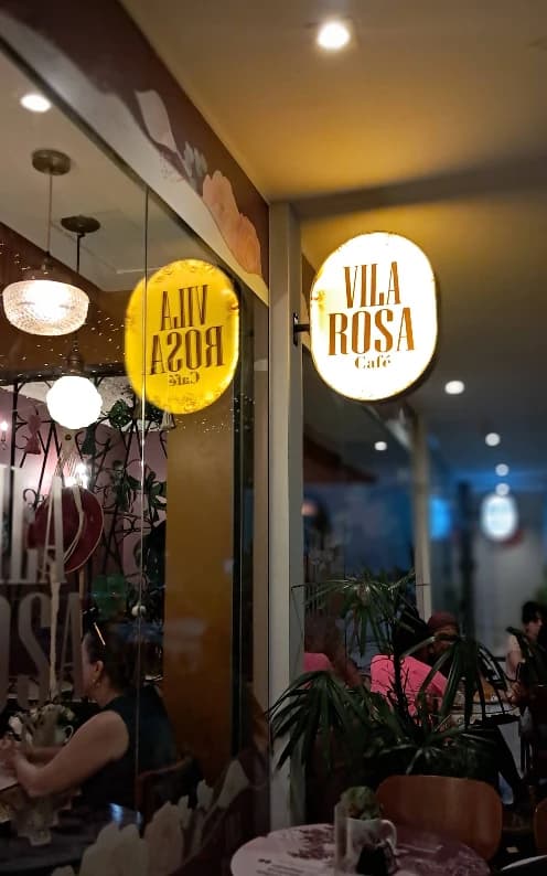 Fachada da Vila Rosa Café à noite, com os dois letreiros circulares iluminados e o salão visto pela vidraça