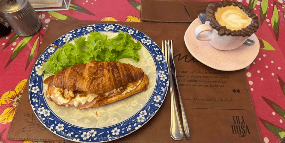 Croissant recheado e cappuccino sobre prato azul floral e toalha estampada, na Vila Rosa Café