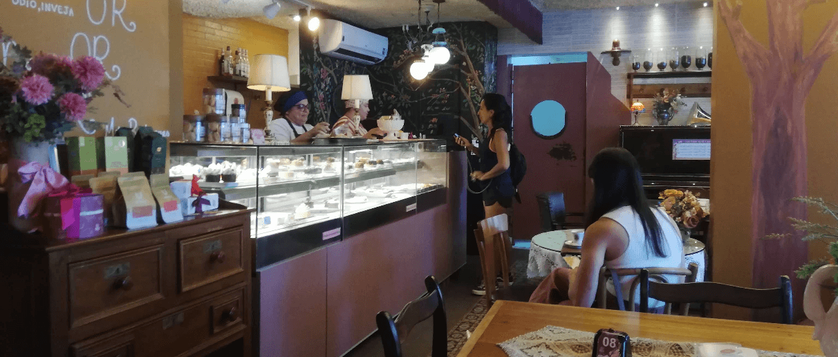 Salão da Vila Rosa Café com a vitrine de doces, equipe atrás do balcão, mural de folhagens e o piano ao fundo