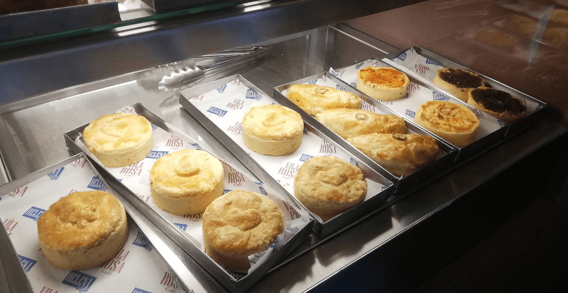 Empadas e quiches recém-assadas em bandejas sobre papéis com a marca da Vila Rosa Café